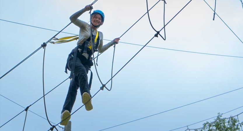 A person wearing safety gear is secured by ropes as they balance on an obstacle of a high ropes course. 